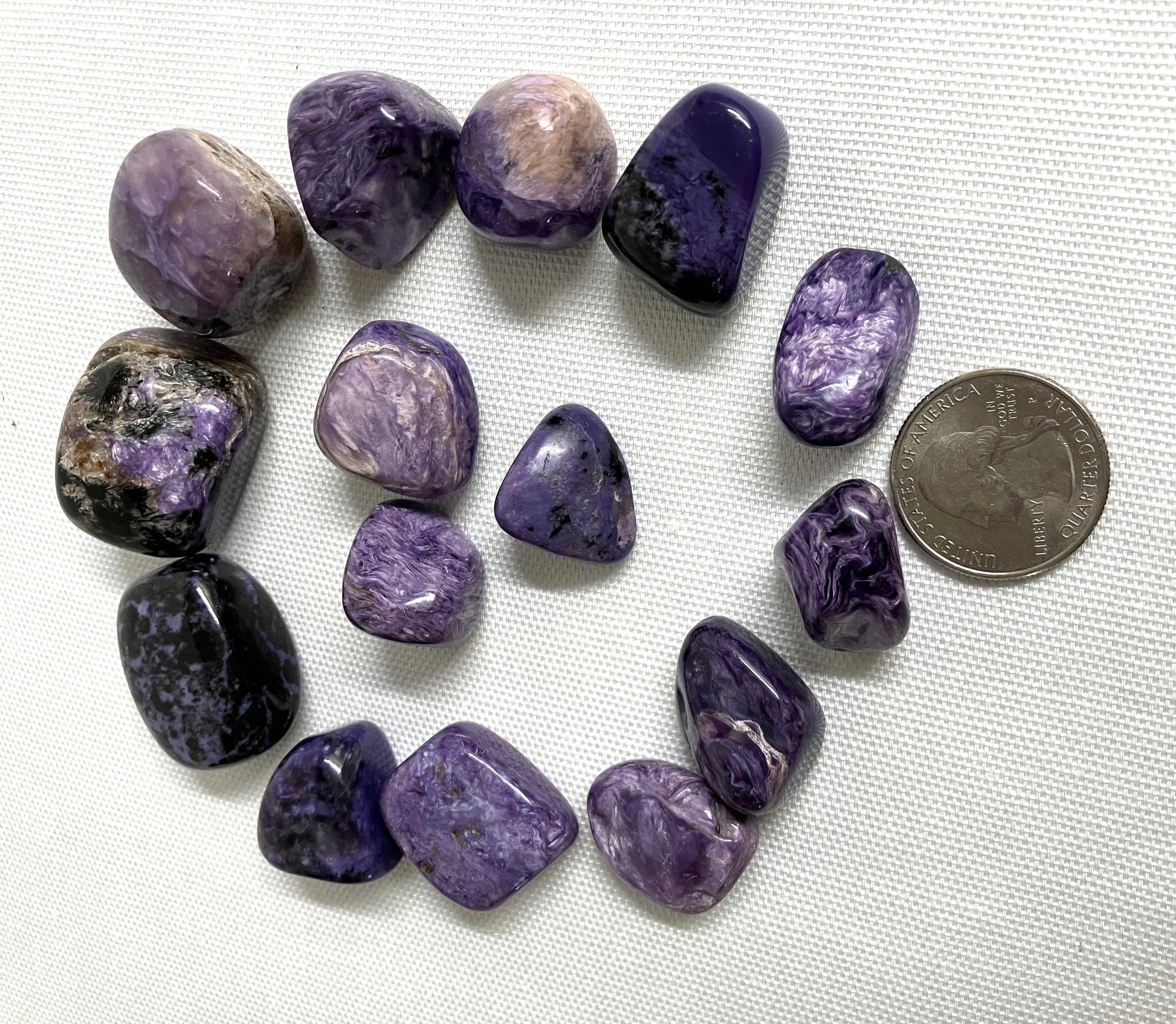 Charoite Tumbled Crystals arranged in a circle on a white fabric background with a quarter for scale.