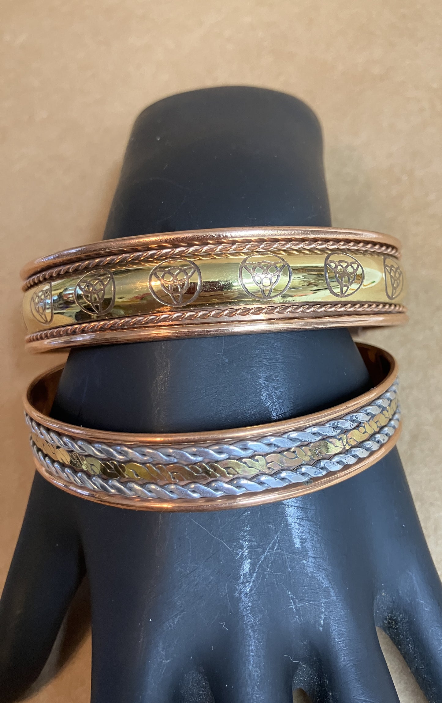 Two copper bracelets with intricate designs on a black stand against a beige background