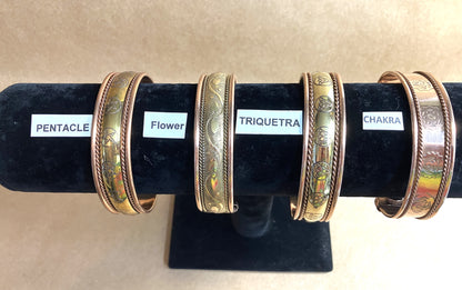 Four Copper Magnetic Cuff Bracelets  on a black stand with labels indicating 'Pentacle', 'Flower', 'Triquetra', and 'Chakra'.